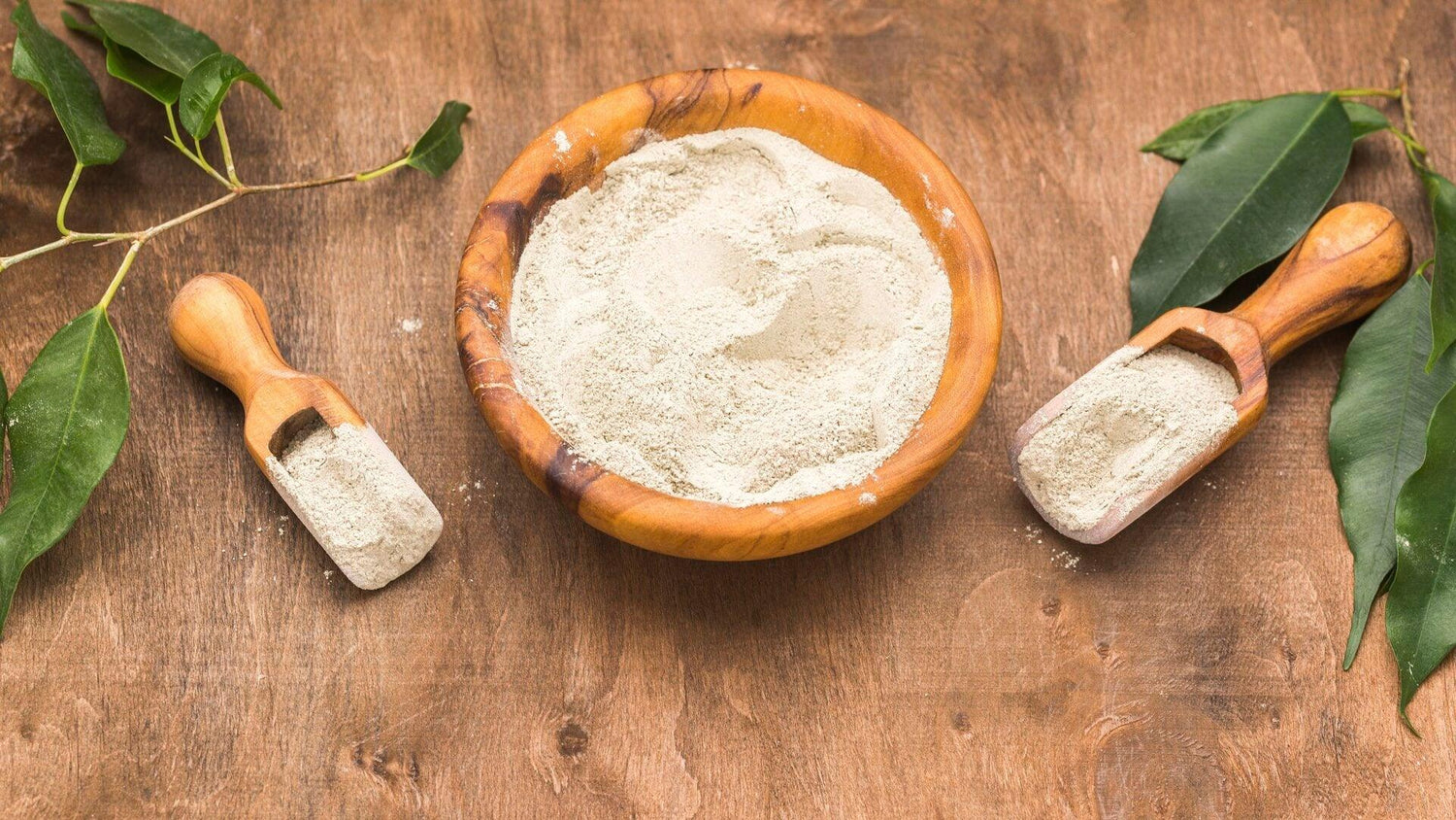 Wooden bowl and scoops filled with white fine powder on wooden table with green leaves