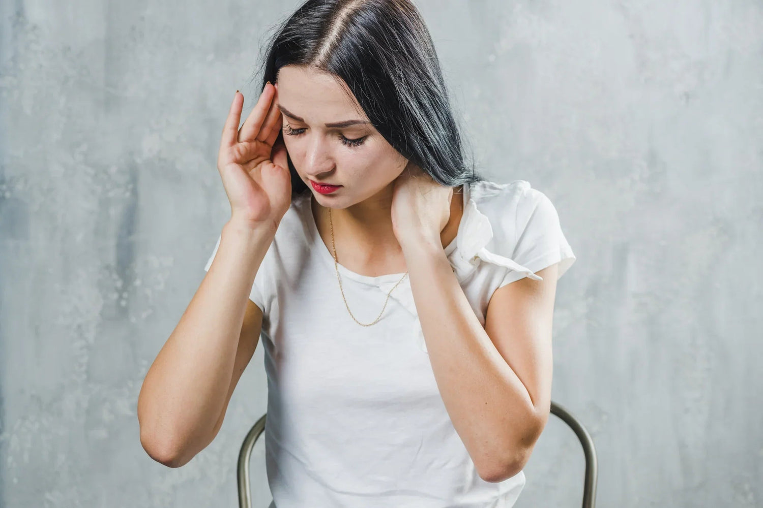 Young woman in white shirt with long dark hair holding head and neck, showing signs of headache